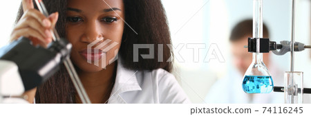An African woman in laboratory with microscope and liquids in flasks working with them An African woman in laboratory with microscope and liquids in flasks working with them 74116245
