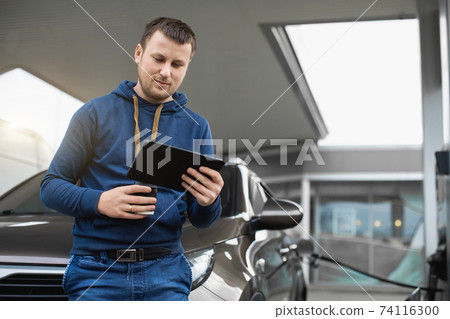 Young handsome Caucasian man, standing in front of his car and working on tablet pc, while refueling the auto at gas station. Blurred petrol station on the background 74116300