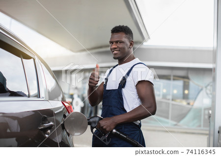 Close up portrait of a happy young African guy, professional gas station worker in uniform, smiling and showing thumb up to camera, while refueling modern car 74116345