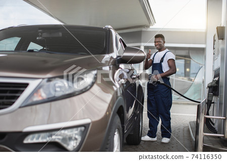Gas station, transportation and energy concept. African man, professional gas station worker in uniform, smiling and showing thumb up to camera, while refueling modern car Gas station, transportation and energy concept. African man, professional gas station worker in uniform, smiling and showing thumb up to camera, while refueling modern car 74116350