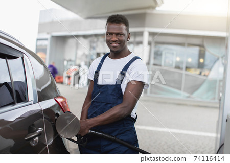 Portrait of a smiling African American gas station worker in workwear, refueling luxury car with gasoline at the station. Car refueling at petrol station Portrait of a smiling African American gas station worker in workwear, refueling luxury car with gasoline at the station. Car refueling at petrol station 74116414