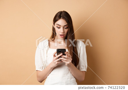 Woman look surprised at smartphone screen, reading shocking message on cell phone, standing amazed on beige background 74122320