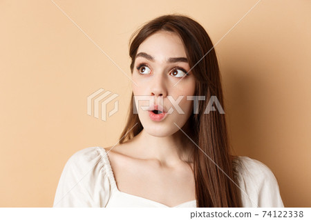 Close up portrait of excited pretty woman looking aside logo, say wow with amazed face, standing on beige background 74122338