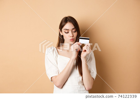 Cute young woman looking thoughtful at plastic credit card, thinking of shopping, standing on beige background 74122339