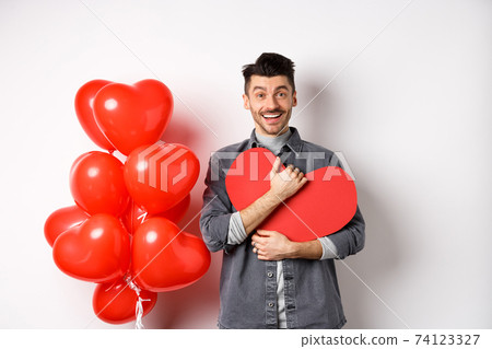 Young romantic man looking with love at camera, hugging Valentine heart card and smiling happy, celebrating lovers holiday, standing near red balloons, white background 74123327
