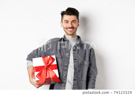 Happy Valentines day. Smiling handsome man bring gift on romantic date, holding box with present and looking romantic at camera, white background Happy Valentines day. Smiling handsome man bring gift on romantic date, holding box with present and looking romantic at camera, white background 74123329