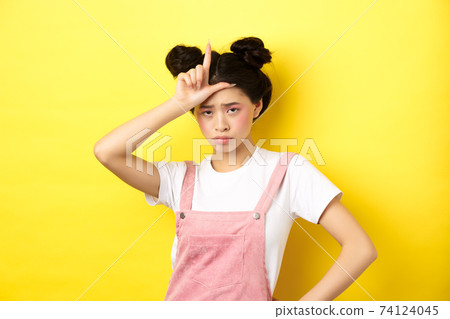 Sad girl showing loser sign on forehead and sulking upset, feeling disappointed in herself, standing on yellow background 74124045