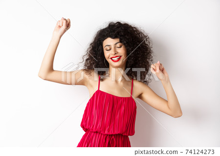 Carefree woman with curly hair, dancing at party, wearing red dress, relaxing with music, standing on white background 74124273