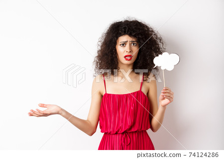 Confused young woman in red dress, frowning and looking upset, holding comment cloud stick, standing against white background 74124286