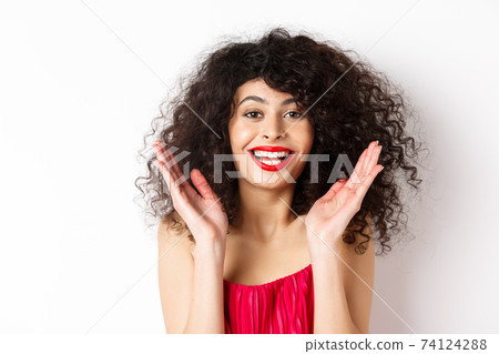 Close-up of fashionable woman with curly hair and red dress, clap hands smiling happy, applause you, standing on white background 74124288