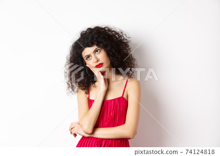 Annoyed and bored young woman with curly hair, look away distressed, lean face on hand, standing bothered in red dress on white background 74124818