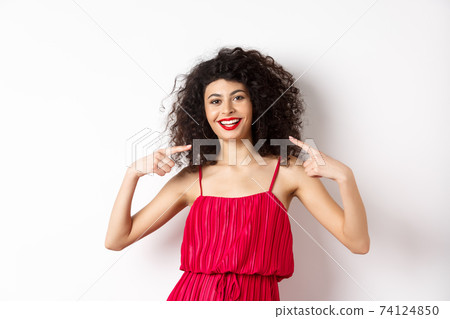 Beautiful woman with curly hairstyle, makeup and red dress, pointing at herself, smiling with white teeth, standing on white background Beautiful woman with curly hairstyle, makeup and red dress, pointing at herself, smiling with white teeth, standing on white background 74124850