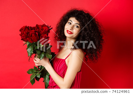 Happy beautiful woman in dress, holding flowers and smiling romantic, standing against red background 74124961