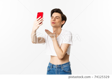 Beautiful gay man in crop top sending air kiss at phone camera, taking selfie or video chat on smartphone, standing over white background Beautiful gay man in crop top sending air kiss at phone camera, taking selfie or video chat on smartphone, standing over white background 74125329
