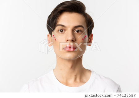 People, lgbtq and beauty concept. Close-up of beautiful androgynous man with glitter on face, looking at camera, standing over white background 74125587