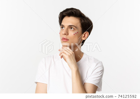 People, lgbtq community and lifestyle concept. Beautiful androgynous man with glitter on face, looking at upper left corner sensual, standing over white background 74125592
