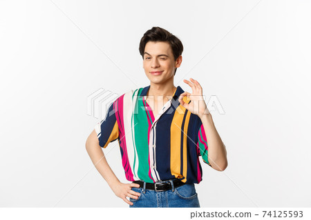 Confident young androgynous man showing okay sign, approve and agree, smiling and winking at camera, guarantee and recommend, standing over white background 74125593
