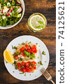 Halibut fish fillet baked with tomatoes on white plate. Salad with radish and cucumber in bowl. Wooden rustic table, top view 74125621