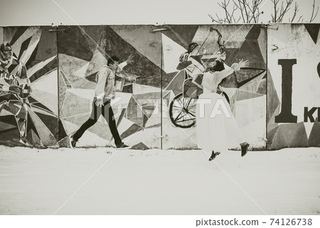 Khmelnytskyi, Ukraine - 27.11.2018: newlyweds in wedding clothes and denim jackets jumping against wall pop art street in winter 74126738