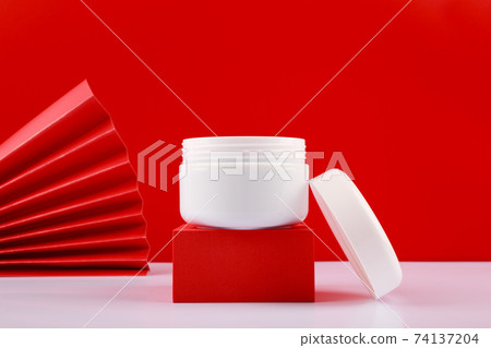 Selective focus, close up of opened jar with cream or mask on red pedestal against red background decorated with waver 74137204