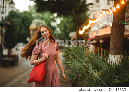 Young beautiful red-haired long-haired woman in a summer pink dress walks through the streets of a summer city. 74152885