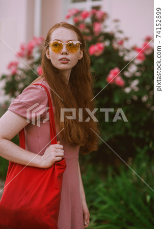 Young beautiful red-haired long-haired woman in a summer pink dress walks through the streets of a summer city. 74152899