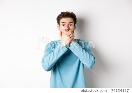 Image of shocked young man staring startled and covering mouth with hands, hear gossips, standing on white background Image of shocked young man staring startled and covering mouth with hands, hear gossips, standing on white background 74154127