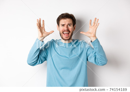 Image of freaked out young man screaming and shaking hands, shouting at camera frustrated or worried, panicking over white background 74154189