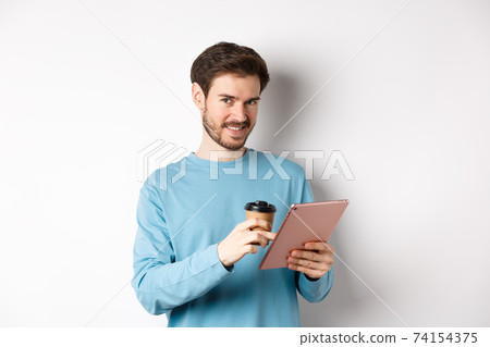 Handsome bearded man smiling at camera, drinking coffee and reading on digital tablet, standing over white background Handsome bearded man smiling at camera, drinking coffee and reading on digital tablet, standing over white background 74154375