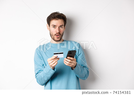 E-commerce and shopping concept. Excited man looking amused while making order online, holding plastic credit card and smartphone, standing on white background E-commerce and shopping concept. Excited man looking amused while making order online, holding plastic credit card and smartphone, standing on white background 74154381