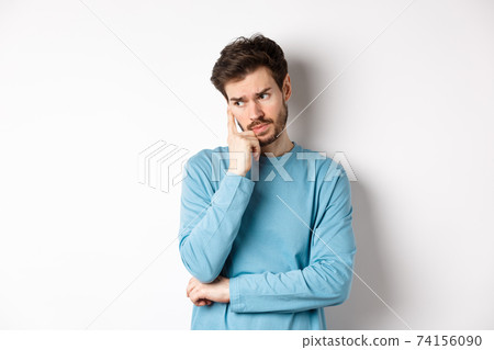 Worried young man with beard in sweatshirt, looking away pensive and thinking, standing troubled against white background 74156090