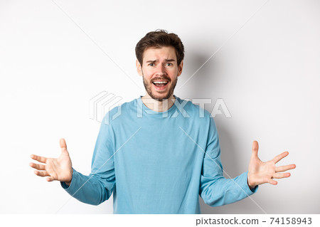 Image of frustrated and worried young man shaking hands, trying to explain something, standing anxious against white background 74158943
