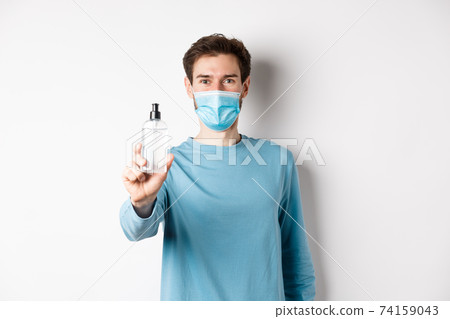 Covid-19, health and quarantine concept. Young man in medical mask stretch out hand and showing sanitizer, recommending antiseptic, standing over white background Covid-19, health and quarantine concept. Young man in medical mask stretch out hand and showing sanitizer, recommending antiseptic, standing over white background 74159043