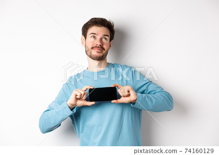 E-commerce and shopping concept. Smiling young man showing mobile screen and dreaming about something, looking at upper right corner nostalgic, standing over white background 74160129