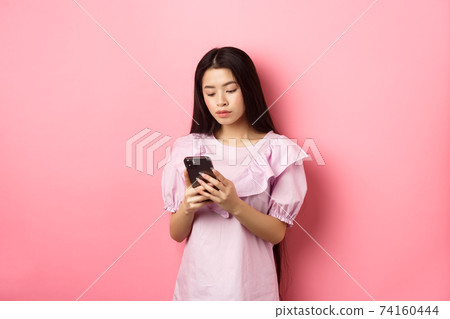 Teenage asian girl chatting on mobile phone, look at smartphone screen with serious face, standing against pink background 74160444