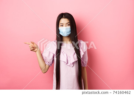 Covid-19, pandemic lifestyle concept. Cute asian teen girl in medical mask pointing finger left at logo, showing advertisement, standing in dress on pink background Covid-19, pandemic lifestyle concept. Cute asian teen girl in medical mask pointing finger left at logo, showing advertisement, standing in dress on pink background 74160462