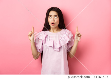 Concerned and sad asian girl frowning, looking and pointing fingers up at bad news, standing against pink background 74161493