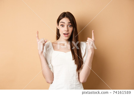 Wow look here. Amazed cute girl making announcement, pointing fingers up and look excited, showing promo offer, beige background 74161589