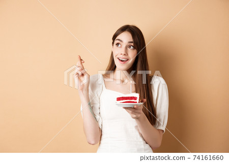 Hopeful birthday girl making wish with fingers crossed, looking up dreamy, holding bday cake with candle, beige background Hopeful birthday girl making wish with fingers crossed, looking up dreamy, holding bday cake with candle, beige background 74161660