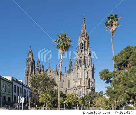 View on beautiful parish church of San Juan Bautista, impressive Neogothic Cathedral in Arucas, Gran Canaria, Spain. Blue sky and palm trees background 74162484
