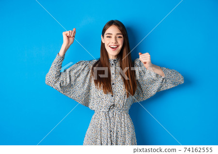 Cheerful young woman in dress raising hands up and smiling excited, triumphing over winning prize, celebrating victory, standing on blue background Cheerful young woman in dress raising hands up and smiling excited, triumphing over winning prize, celebrating victory, standing on blue background 74162555