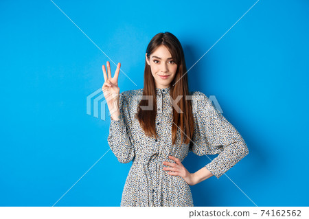 Young beautiful girl in dress with natural long hair, showing number three with fingers and smiling, standing against blue background Young beautiful girl in dress with natural long hair, showing number three with fingers and smiling, standing against blue background 74162562