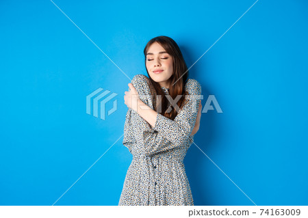 Romantic young natural woman in dress close eyes, smiling and hugging herself with love and tenderness, standing on blue background Romantic young natural woman in dress close eyes, smiling and hugging herself with love and tenderness, standing on blue background 74163009