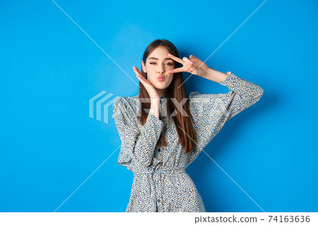 Coquettish young woman in dress showing v-sign on eye and pucker lips for kiss, standing happy on blue background Coquettish young woman in dress showing v-sign on eye and pucker lips for kiss, standing happy on blue background 74163636