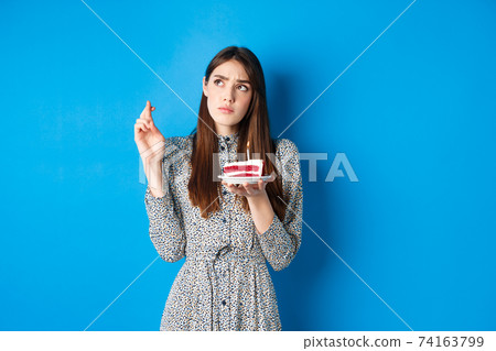 Thoughtful birthday girl making wish, cross fingers good luck, holding cake and looking at upper left corner, blue background Thoughtful birthday girl making wish, cross fingers good luck, holding cake and looking at upper left corner, blue background 74163799
