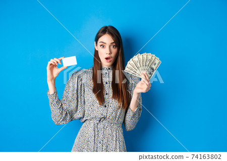 Excited lucky girl showing plastic credit card and money dollar, gasping amazed, buying something expensive, standing on blue background Excited lucky girl showing plastic credit card and money dollar, gasping amazed, buying something expensive, standing on blue background 74163802