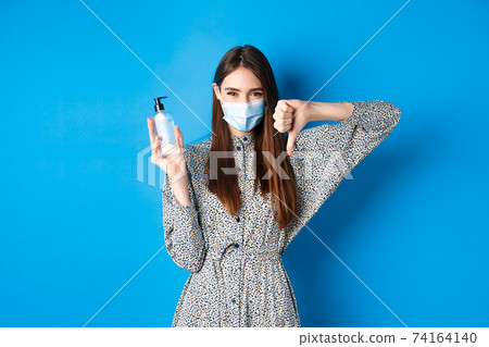 Covid-19, social distancing and healthcare concept. Disappointed girl in medical mask showing bad hand sanitizer and thumbs down, standing on blue background 74164140