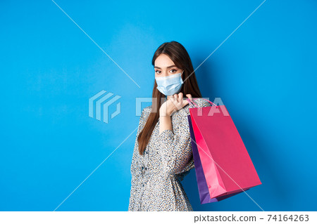 Covid-19, pandemic and lifestyle concept. Elegant woman wear medical mask on shopping, holding bags over shoulder and walking against blue background Covid-19, pandemic and lifestyle concept. Elegant woman wear medical mask on shopping, holding bags over shoulder and walking against blue background 74164263
