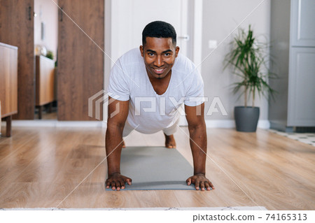 Front view of cheerful African-American man doing push-up on floor at domestic room, looking at camera Front view of cheerful African-American man doing push-up on floor at domestic room, looking at camera 74165313