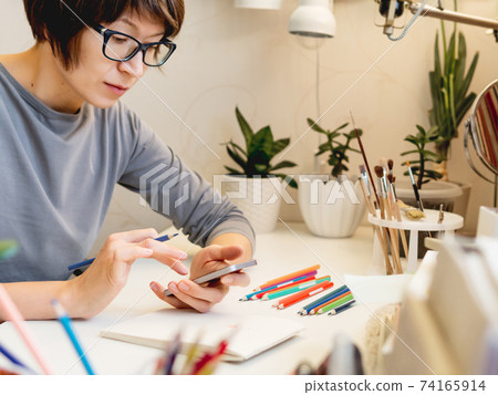 Woman with short hair cut is drawing in notebook with smartphone. Calming hobby, antistress leisure. Artist at work. Cozy workplace. 74165914